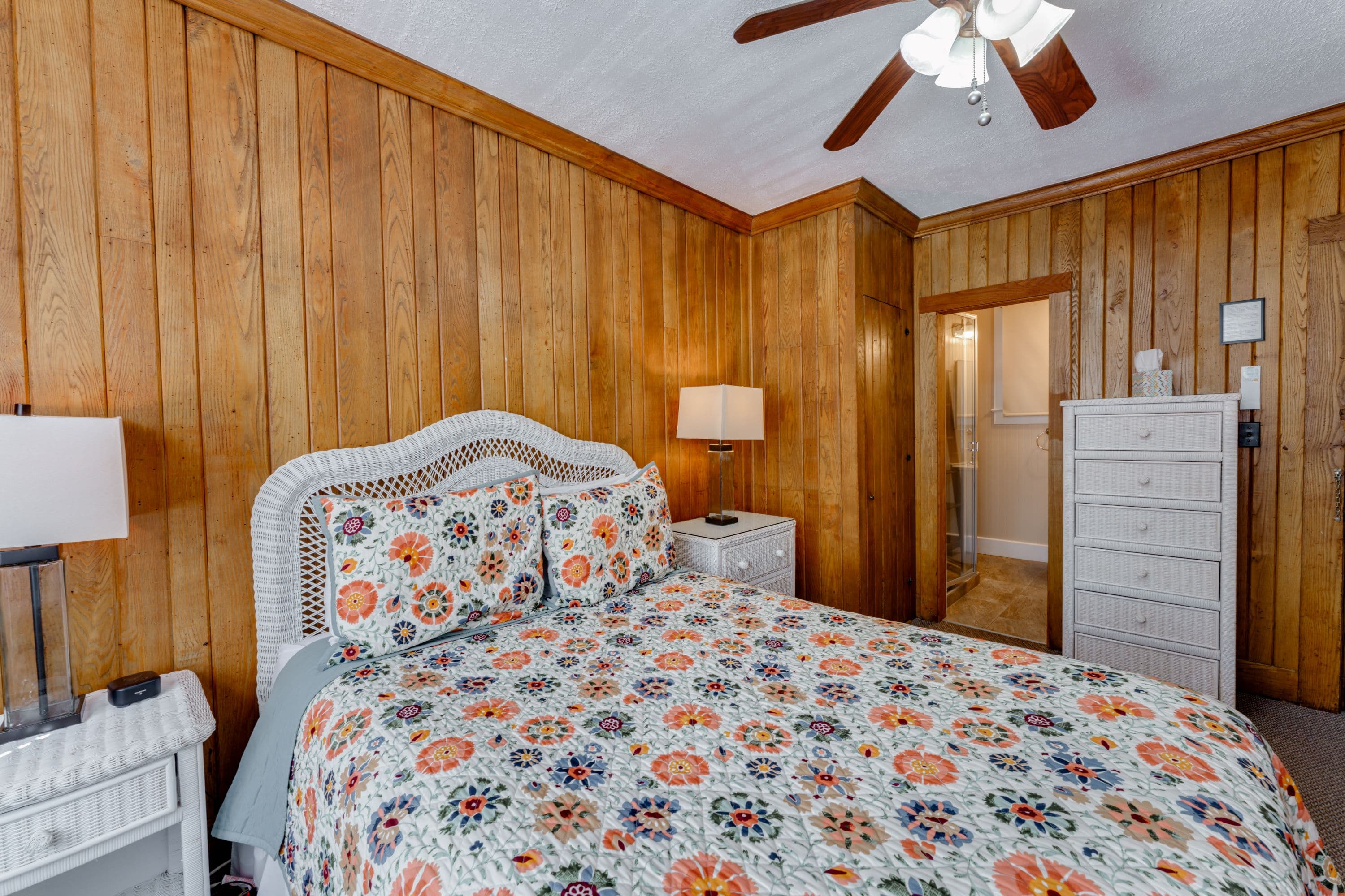 Room features Wormy Chestnut wood paneled walls and white wicker furniture, including night stands, dressers, and a queen bed.