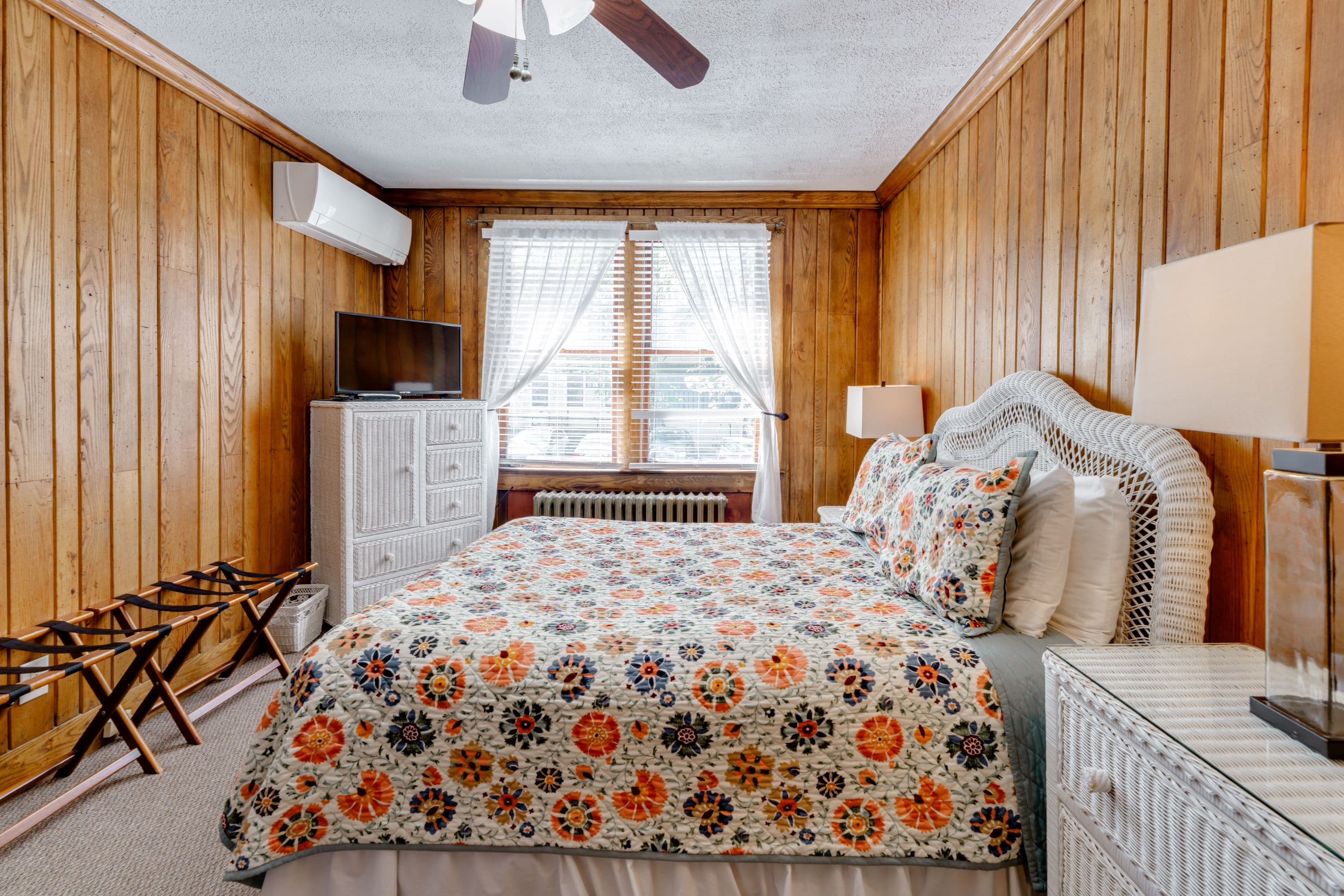 Room features Wormy Chestnut wood paneled walls and white wicker furniture, including night stands, dressers, and a queen bed.