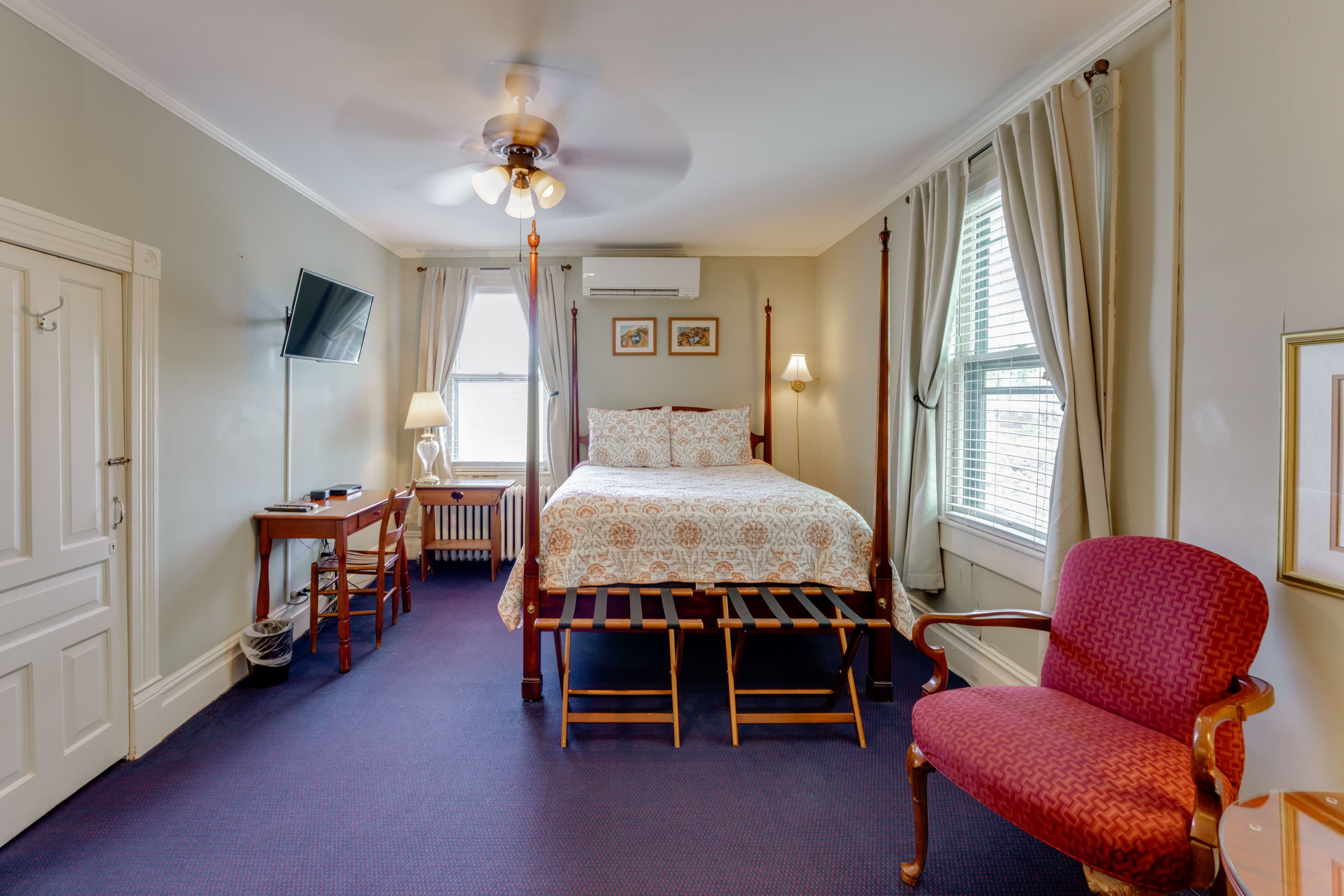 Hepatica Americana Room featuring a four posted queen bed, sitting area with cushioned chairs, a night stand, and a desk and chair with a TV mounted above it.