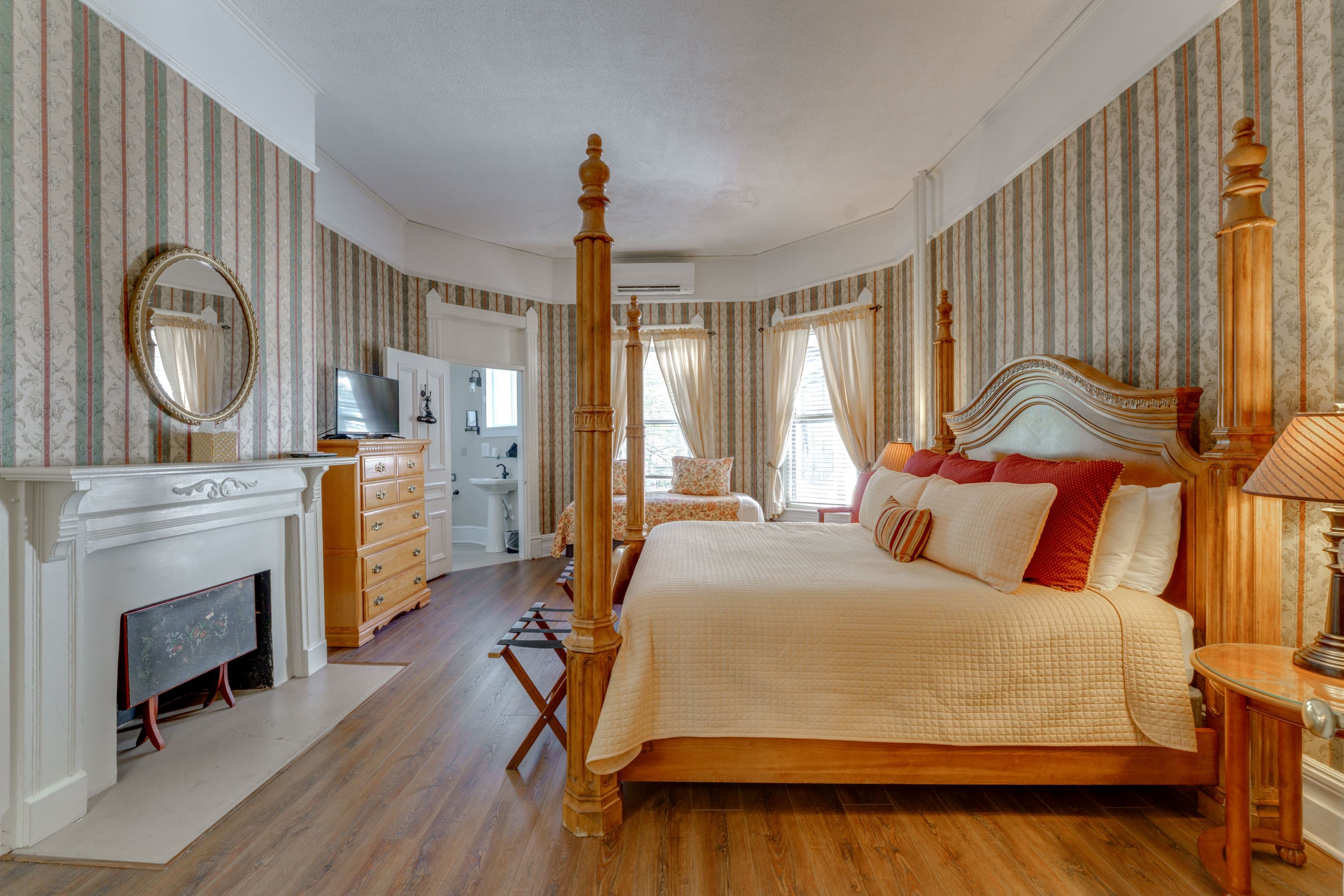 Room features wood flooring, striped wallpaper, a four posted king bed, twin bed, and tall wooden dresser with TV.