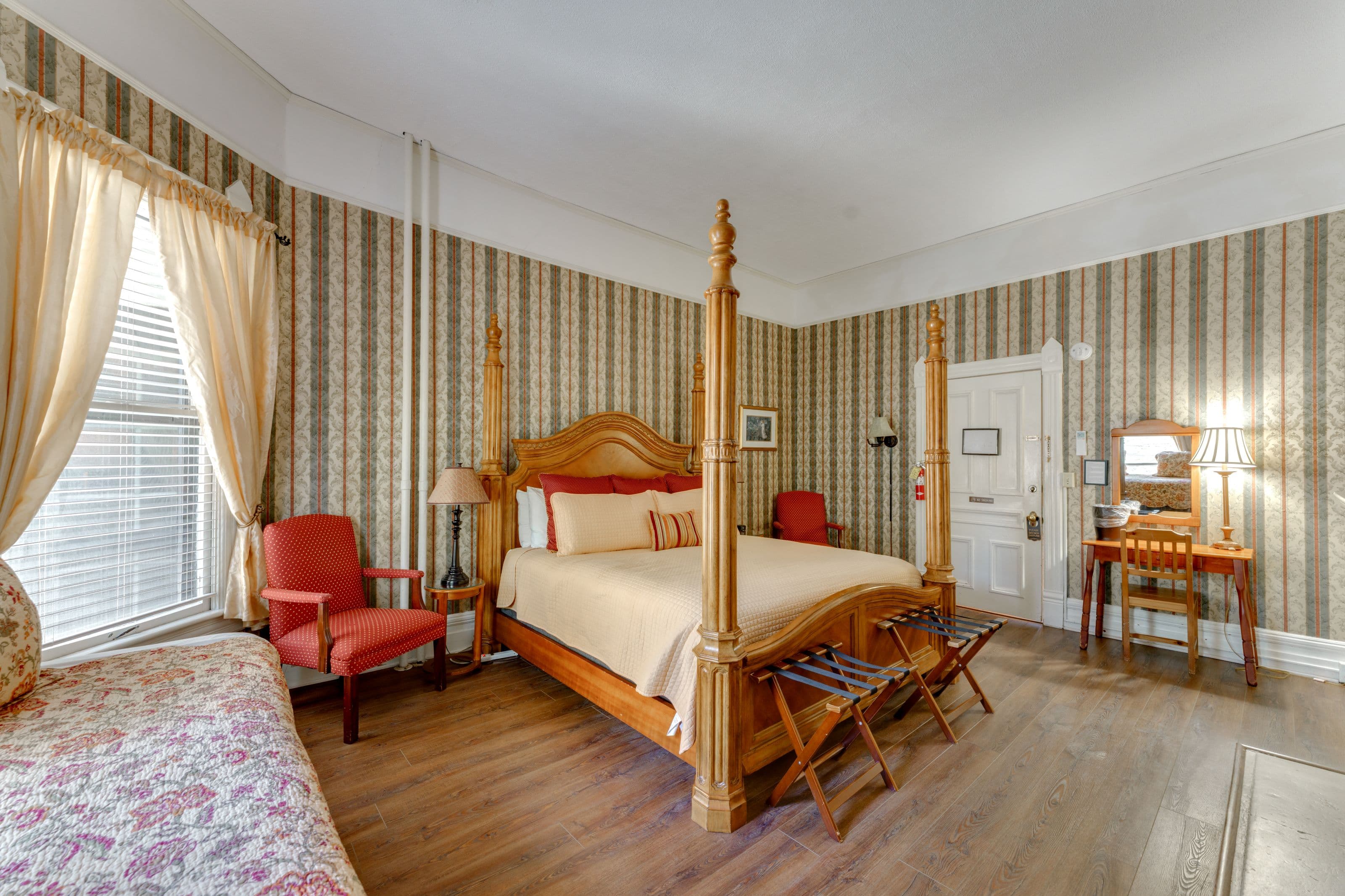 Room features four posted king bed, twin bed, two cushioned chairs, and a desk.