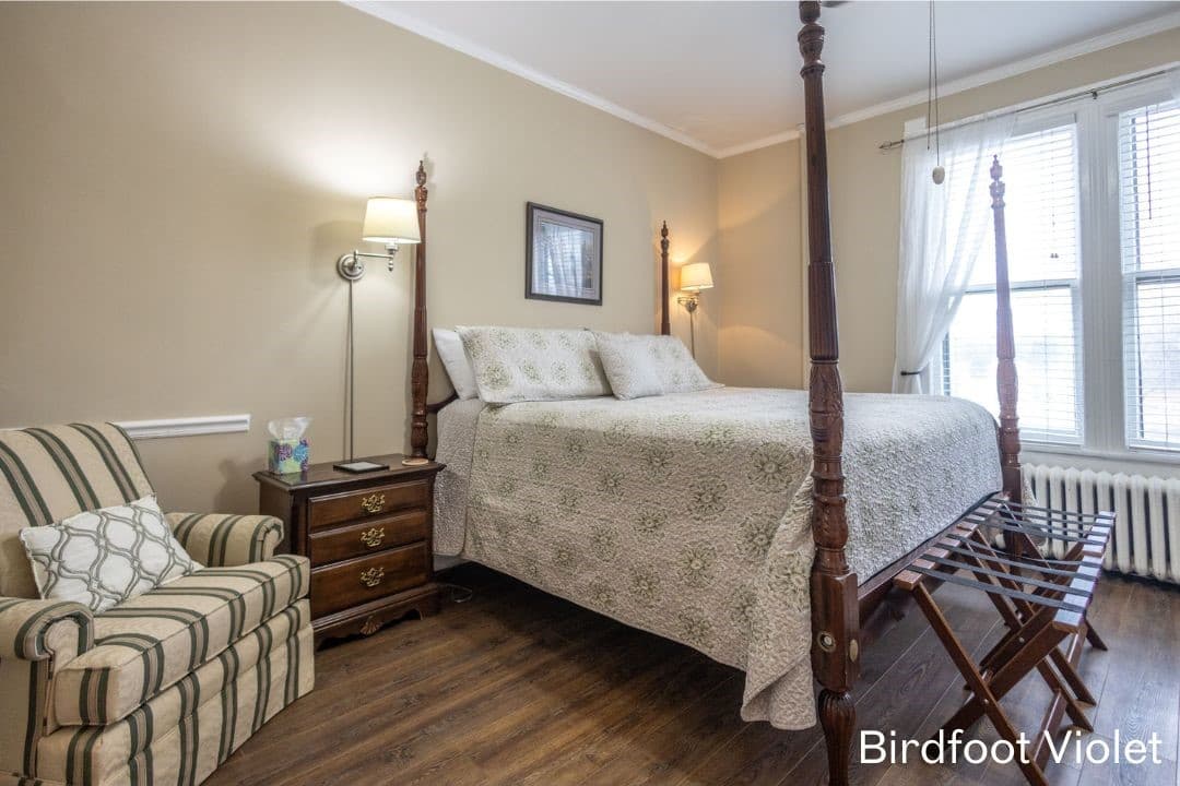 Room features wood flooring, light colored walls, a four posted king bed, and a striped comfy chair.