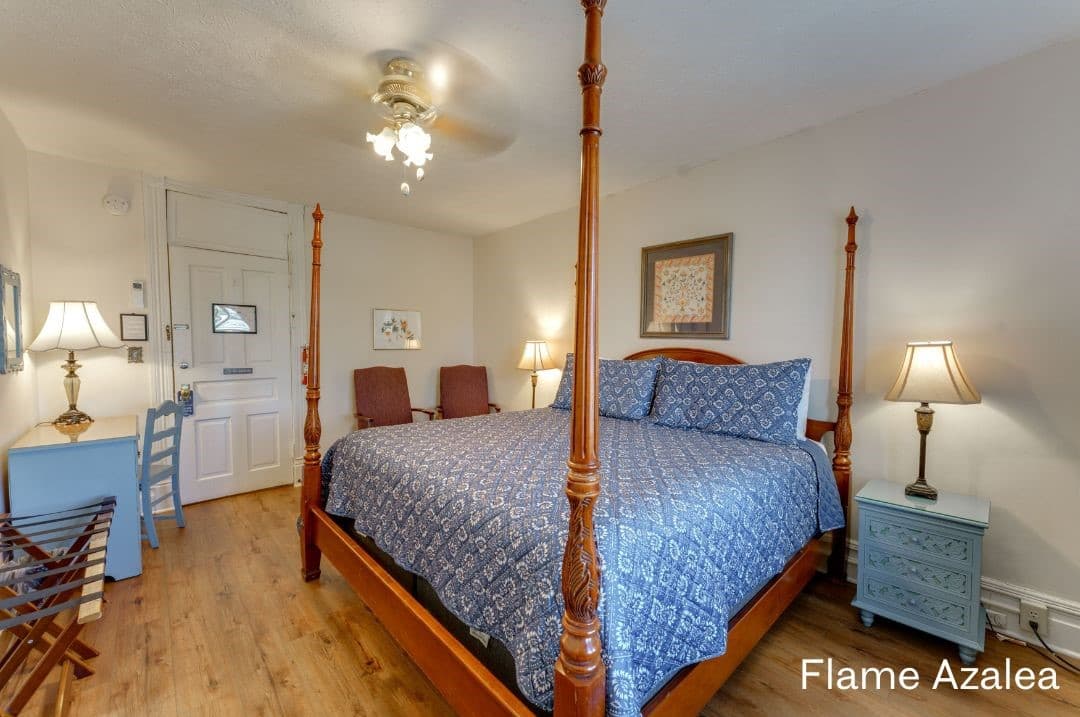 Flame Azalea room features a four-posted king bed, small seating area with cushioned chairs, a desk and chair, a night stand with a lamp on each side, and luggage racks.