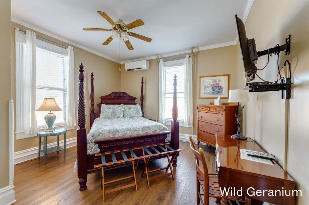Wild Geranium Room featuring a four posted queen bed, round night stand, tall dresser, and desk and chair with a TV mounted above it.