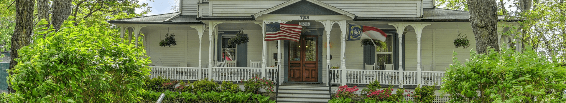 A charming white house with a porch, featuring American and North Carolina flags, surrounded by lush greenery.