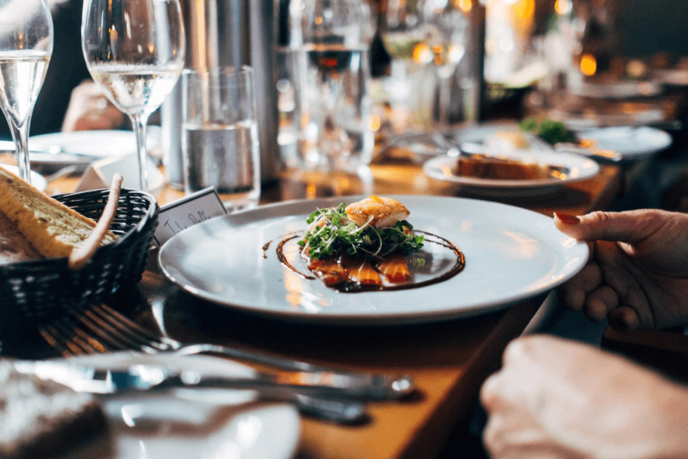 A beautifully plated dish with greens and sauce on a dinner table surrounded by wine glasses and cutlery.