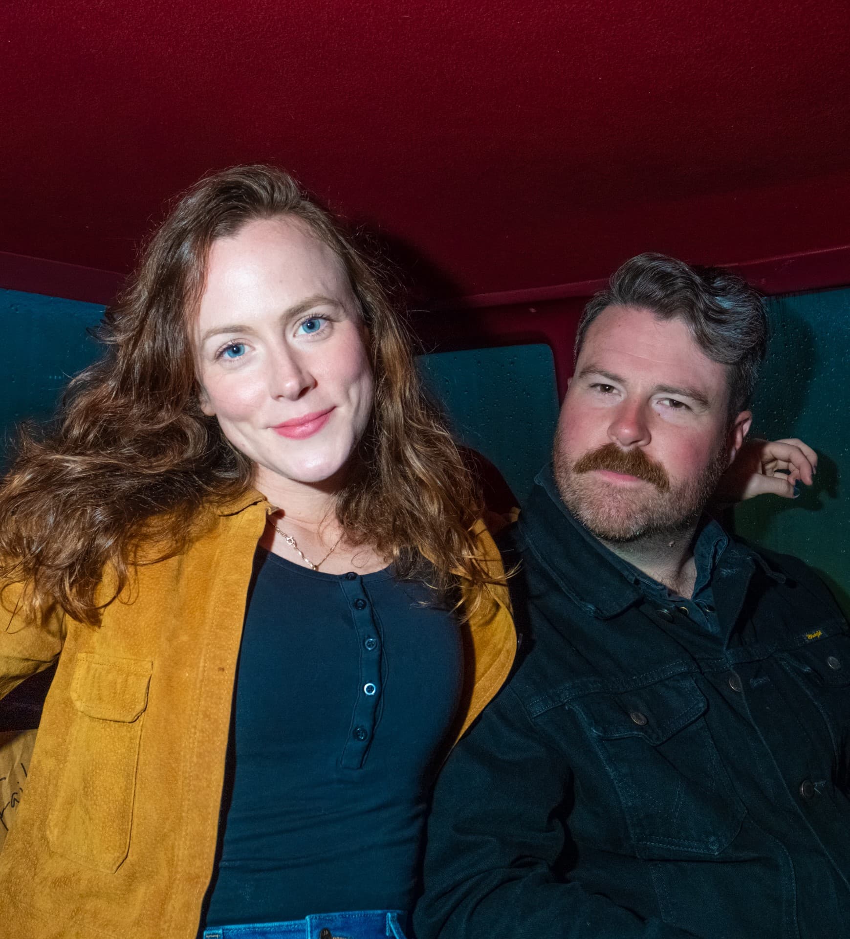 A woman with wavy hair and a gold jacket sits next to a man with a mustache in a cozy setting.