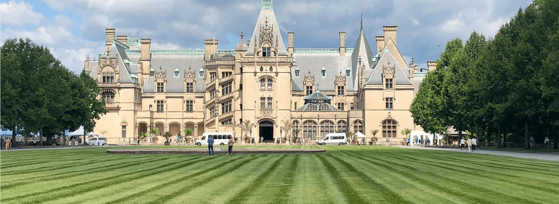 A grand mansion surrounded by neatly striped lawns and trees under a partly cloudy sky.