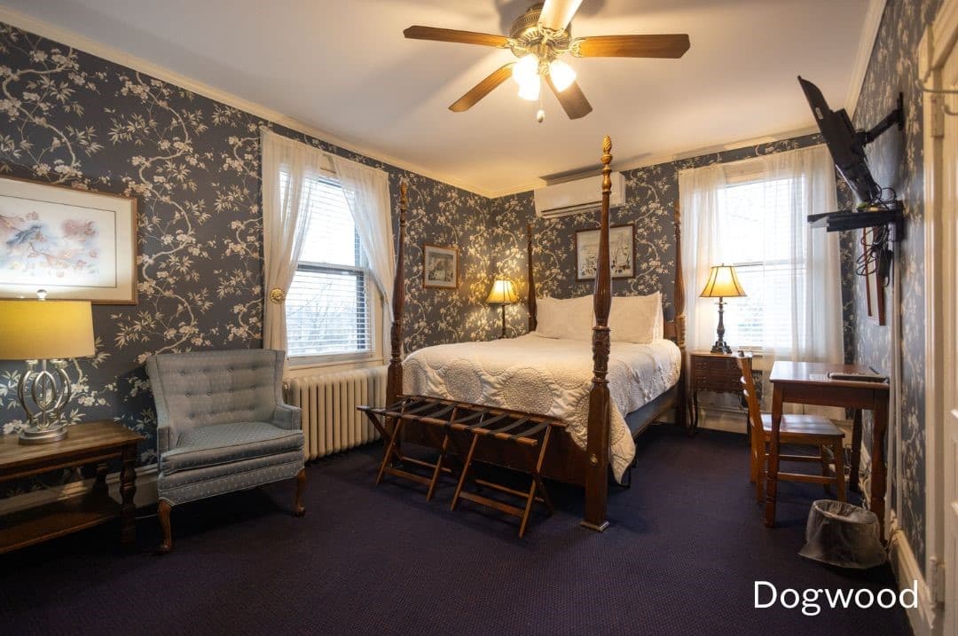 Dogwood Room featuring a four posted queen bed, a night stand on each side, a seating area with cushioned chairs, and a desk and chair with a TV mounted above it.