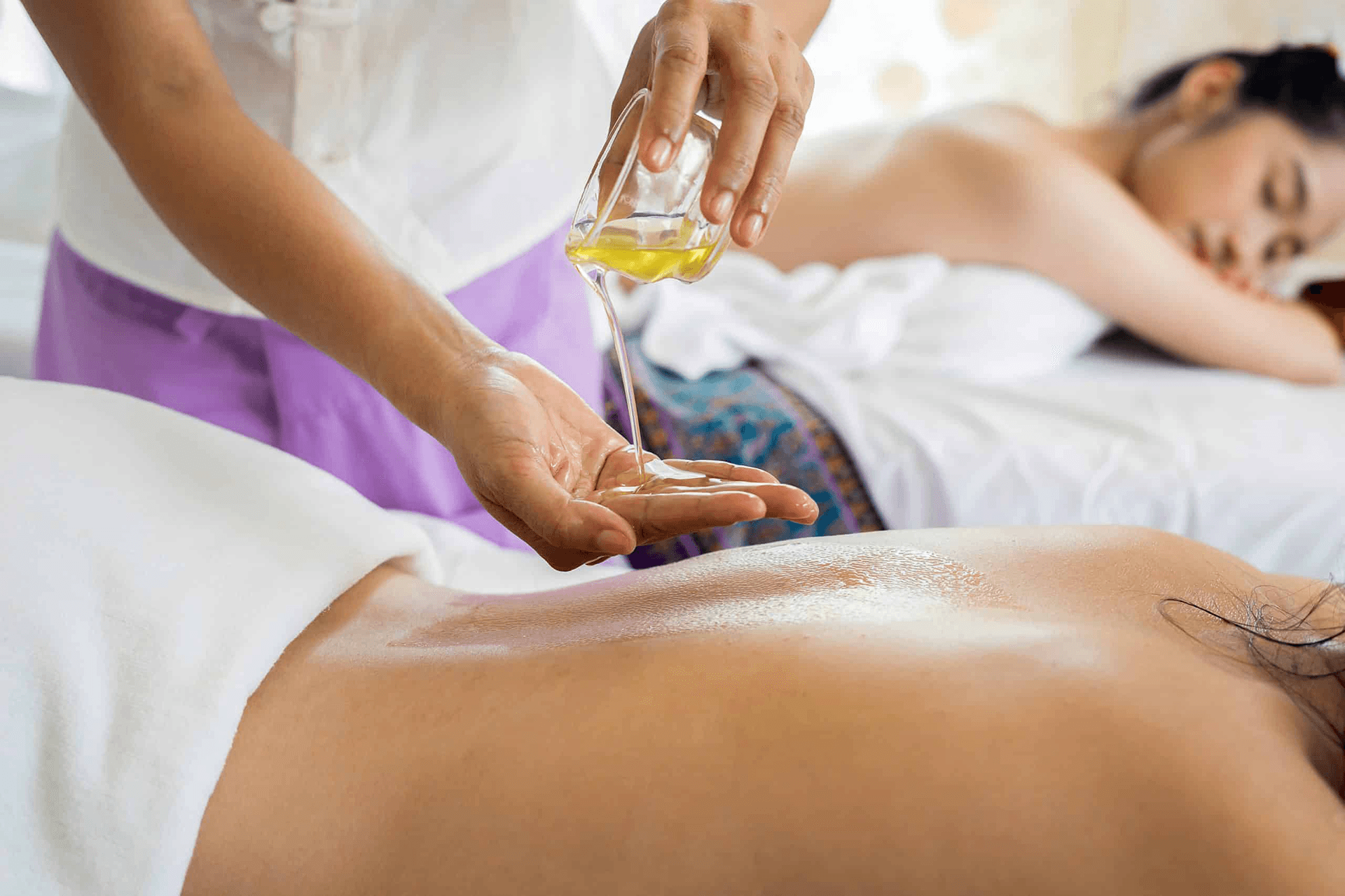 A massage therapist is pouring oil onto a client's back in a tranquil spa setting.