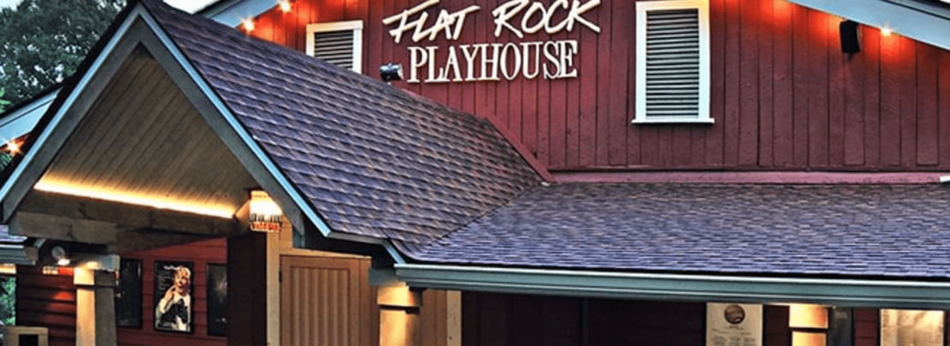 The entrance of Flat Rock Playhouse featuring a well-lit red building and a sloped roof.