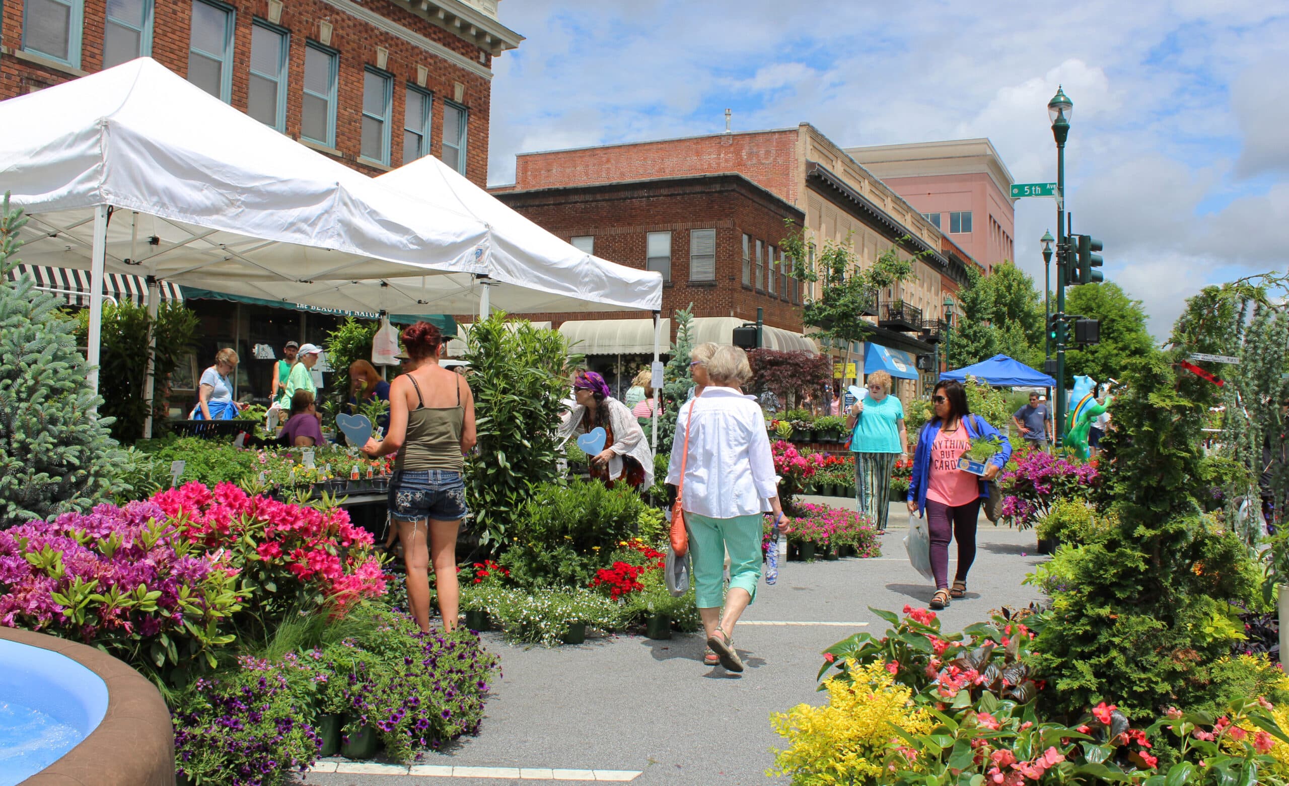 A vibrant farmers market featuring colorful flowers and people browsing various stalls under sunny skies.
