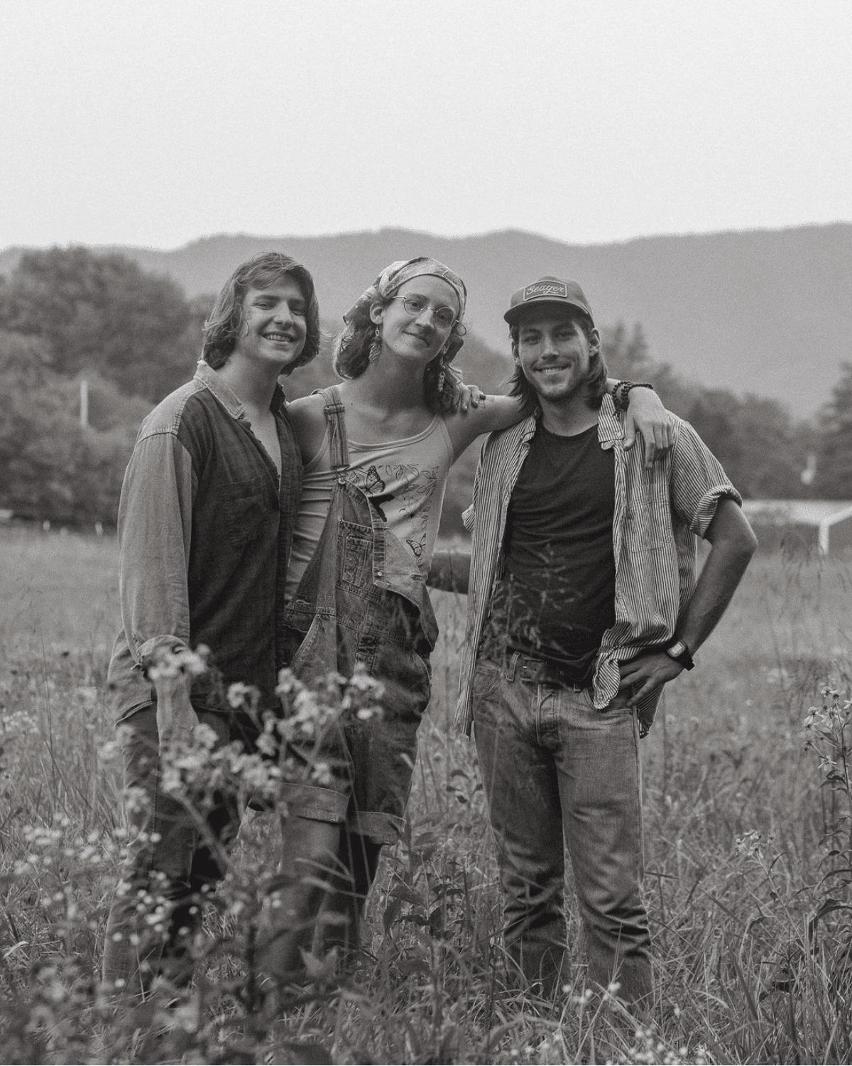 Three individuals posed together in a grassy field with mountains in the background, wearing casual clothing.