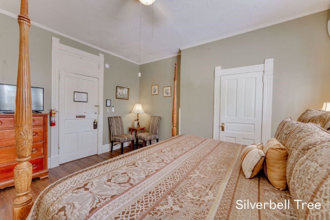 Silverbell Tree room features a four posted king be, small seating area with cushioned chairs, and a tall dresser with a TV on top.