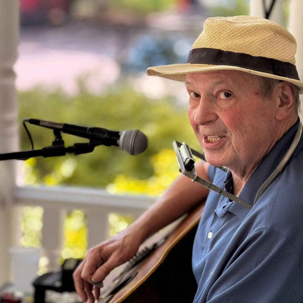 An older man wearing a straw hat plays guitar and smiles at the camera while seated near a microphone.