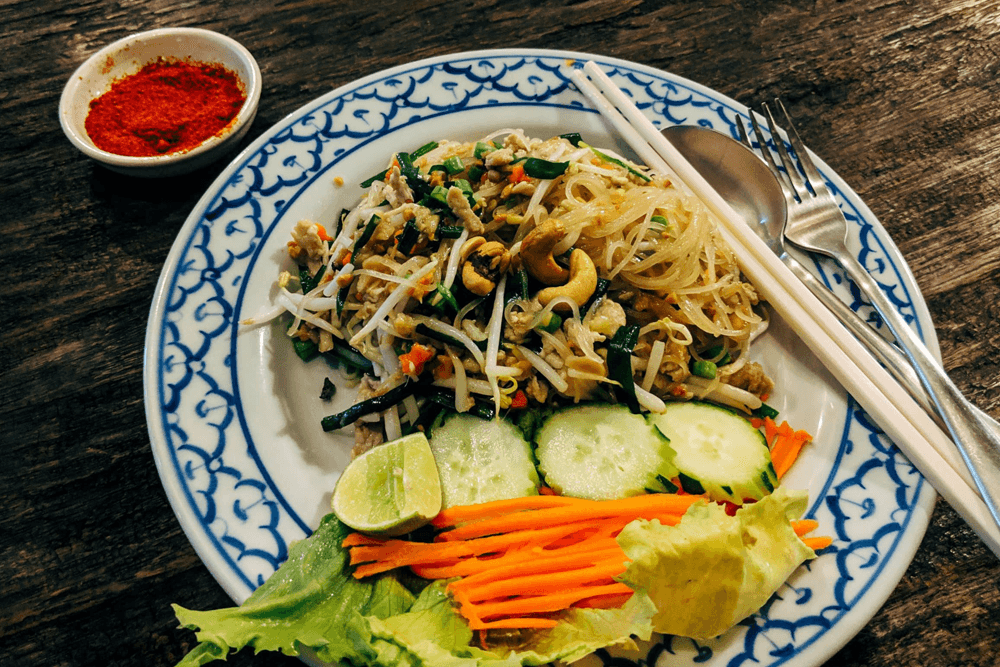 A plate of stir-fried noodles topped with vegetables, cashews, and lime, accompanied by a small bowl of red spice.