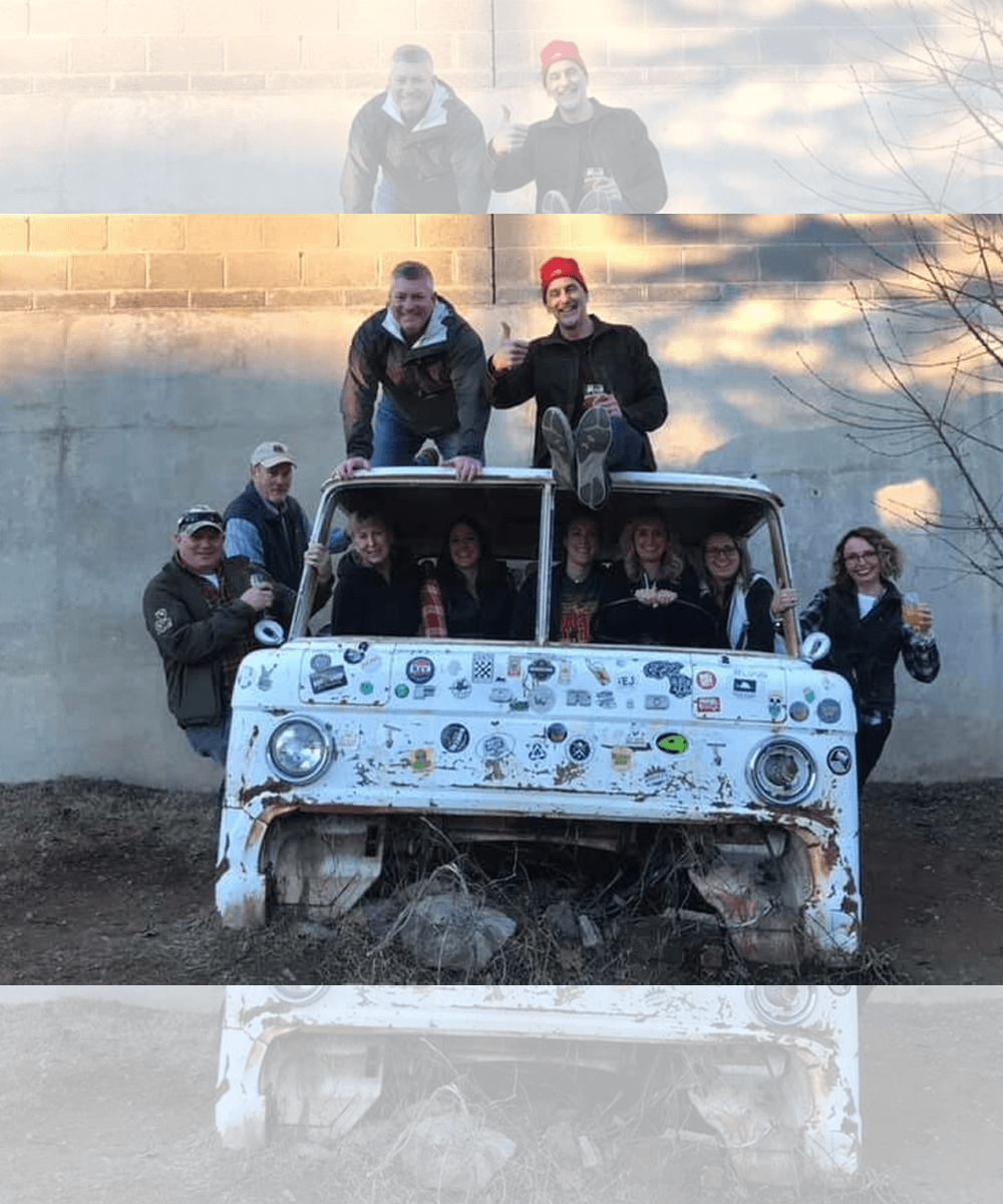A group of eight individuals poses on and around a rusted, sticker-covered vintage vehicle in an outdoor setting.