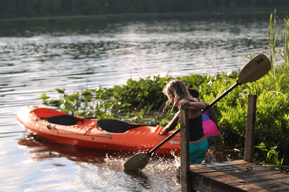A child steps into the water near an orange kayak by a dock.