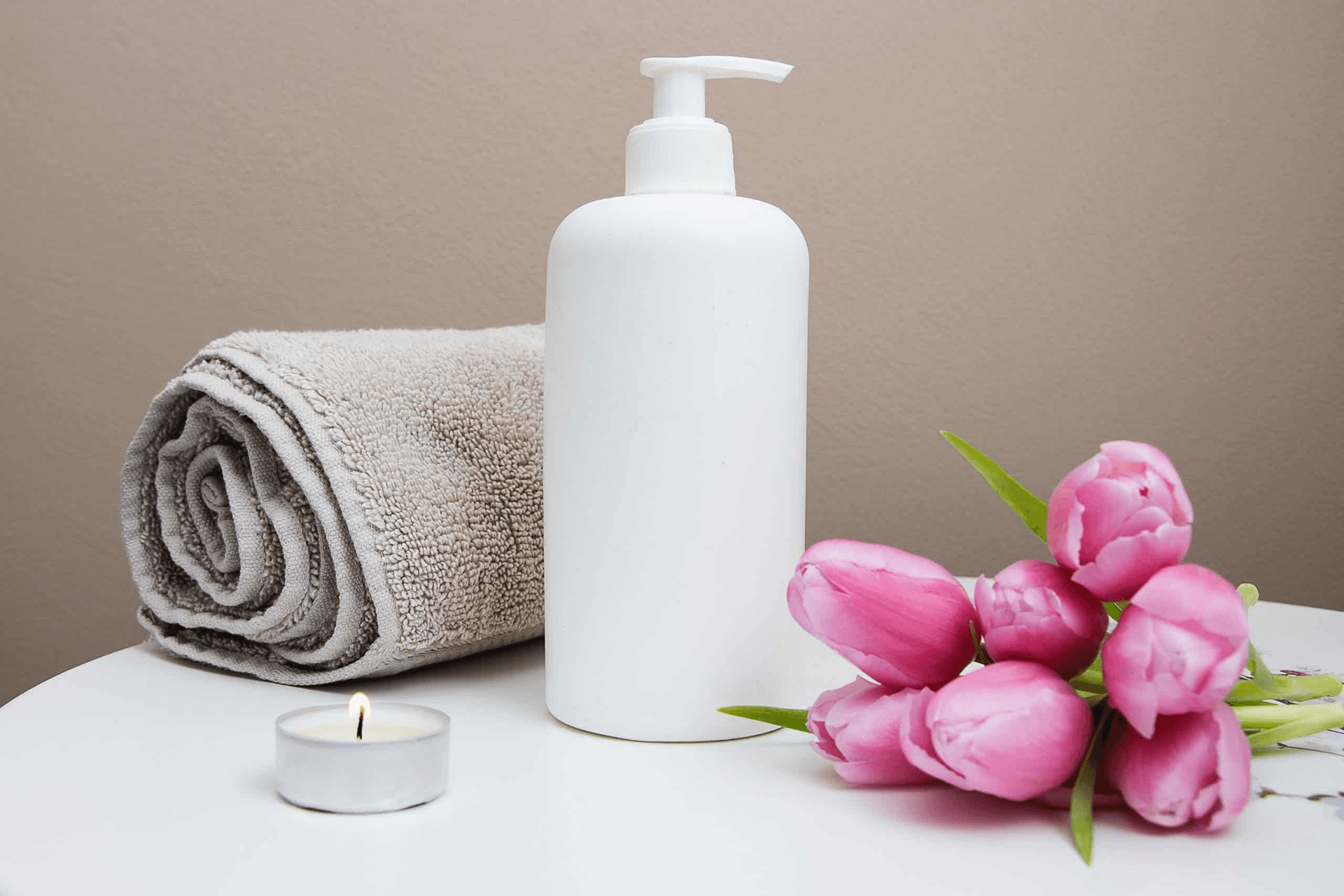 A white lotion bottle beside a rolled towel, a candle, and pink tulips on a table.